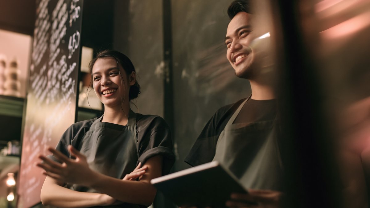 Two café staff in aprons smiling beside a chalkboard menu